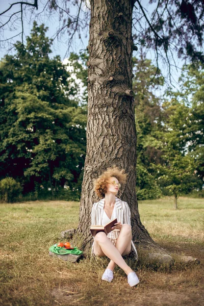 Młoda kobieta siedząca pod drzewem w parku — Zdjęcie stockowe