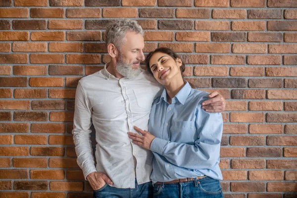 Um homem de cabelos grisalhos abraçando sua esposa enquanto estava perto da parede de tijolos — Fotografia de Stock