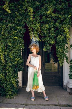 Beyaz uzun elbiseli genç bir kadın elinde yeşil bir ağ torbası tutuyor.