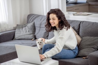 Koyu saçlı genç bir kadın kedisine sarılıyor ve bir video görüşmesi yapıyor.