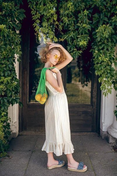 하얀 긴 드레스를 입고 녹색 그물주머니를 손에 들고 선글라스를 쓴 예쁜 여성 — 스톡 사진