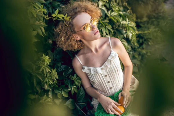 Mujer bonita en un vestido largo blanco y gafas de sol sosteniendo una bolsa de malla verde en sus manos — Foto de Stock