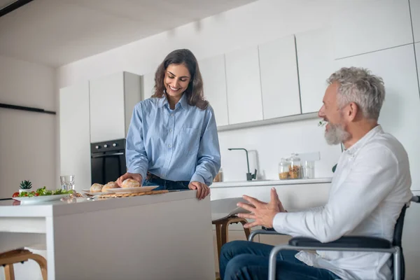 Mujer joven haciendo el desayuno, su marido discapacitado sentado cerca — Foto de Stock