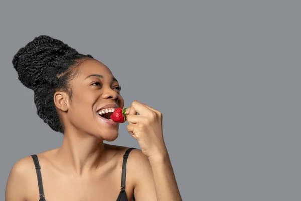 Mulher bonita com pão de cabelo em roupas pretas comendo morango e sorrindo — Fotografia de Stock