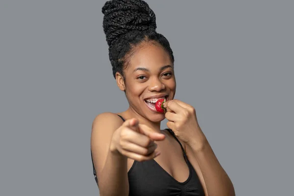 Pretty woman with hair bun in black clothes eating strawberry and smiling — Stock Photo, Image