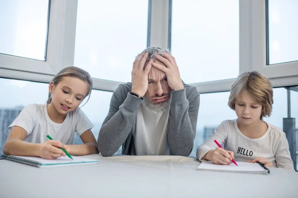 Hombre barbudo de pelo gris sentado con sus hijos en la mesa mientras hacen clases —  Fotos de Stock