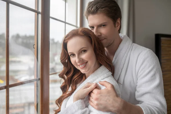 A man and a woman in white bathrobes standing near the window and feeling in love — Stock Photo, Image