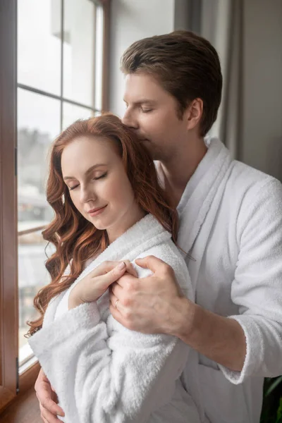 En man och en kvinna i vita badrockar står nära fönstret och känner sig förälskad — Stockfoto