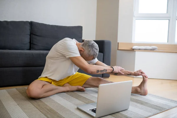 Un hombre maduro en camiseta blanca sentado en el suelo y haciendo yoga en internet — Foto de Stock