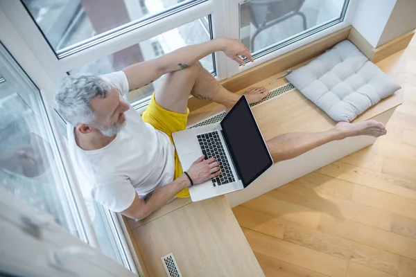 Um homem com uma tshirt branca sentado com um laptop e trabalhando — Fotografia de Stock