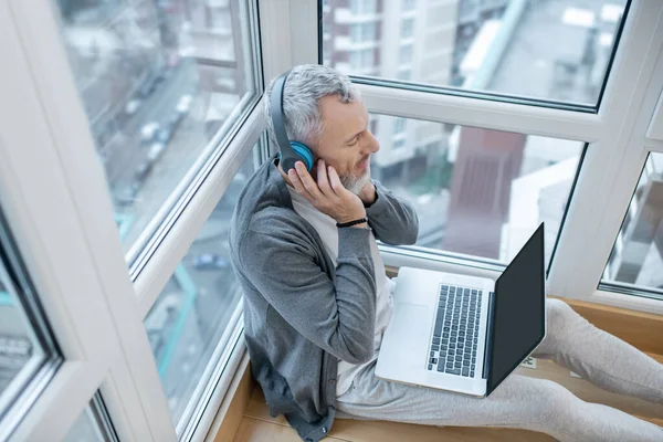 Un hombre maduro con un portátil y en auriculares escuchando algo en Internet —  Fotos de Stock