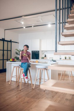 Koyu tenli bir kadın mutfakta oturuyor ve salata yiyor.