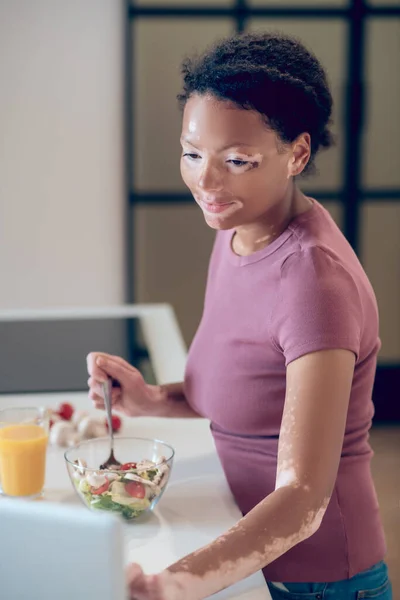 En mörkhyad kvinna äter sallad och tittar på något på laptop — Stockfoto