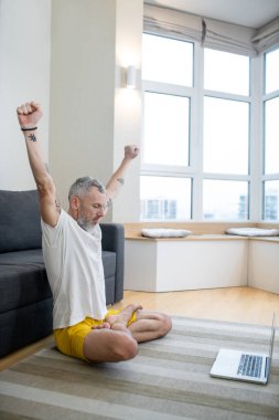 Beyaz tişörtlü olgun bir adam yerde oturuyor ve internetten yoga dersi kaydediyor.