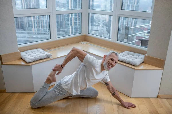 Hombre de pelo gris con una camiseta blanca haciendo yoga y luciendo involucrado — Foto de Stock