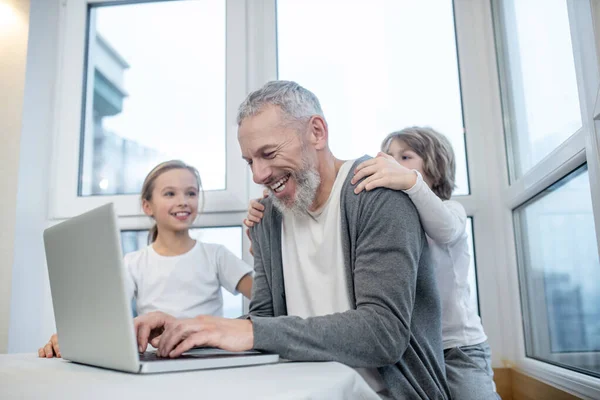 Hombre barbudo de pelo gris que trabaja en el portátil mientras sus hijos lo interfieren —  Fotos de Stock