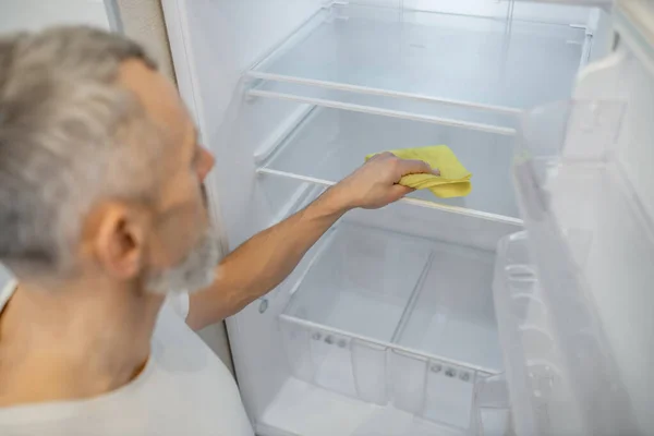 Šedovlasý muž uklízí ledničku v kuchyni. — Stock fotografie