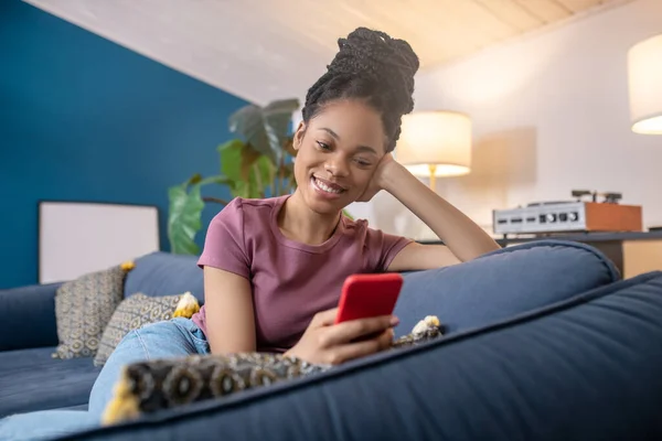 一个拿着智能手机坐在沙发上的年轻的非洲女人 — 图库照片