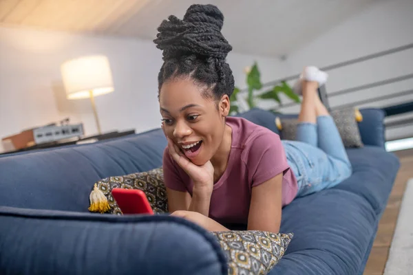 Enthusiastische Afroamerikanerin schaut auf dem Sofa aufs Smartphone — Stockfoto