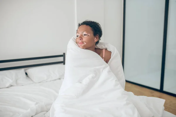 Молодая женщина, сидящая в постели, покрытая одеялом и расслабленная — стоковое фото