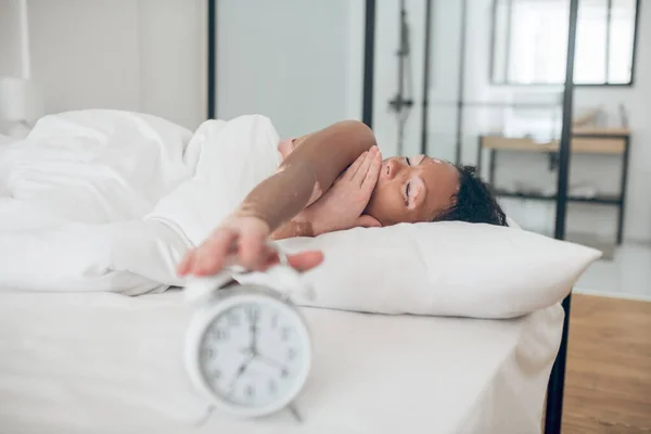 Молода темношкіра жінка лежить у ліжку і вимикає будильник — стокове фото