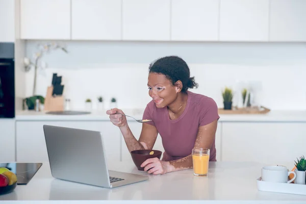 Темнокожая молодая женщина завтракает дома и смотрит что-то на ноутбуке — стоковое фото
