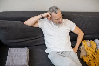 Kanepede oturan gri saçlı bir adam ve yanında yatan kıyafetlere bakıyor.