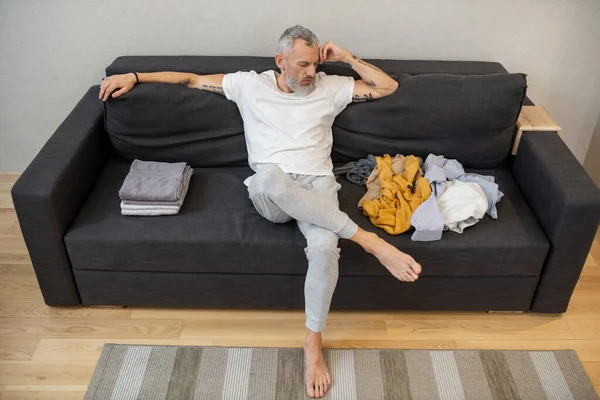 Un hombre de pelo gris sentado en el sofá y mirando la ropa que está cerca — Foto de Stock