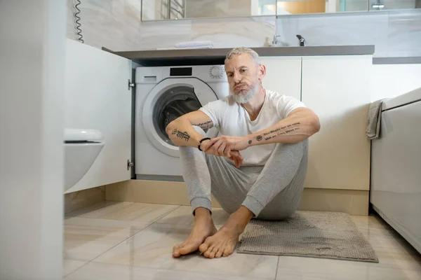 Šedovlasý muž sedí na podlaze v koupelně a vypadá rozrušeně. — Stock fotografie