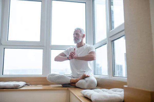 Hombre de pelo gris con una camiseta blanca haciendo yoga y buscando concentrado — Foto de Stock