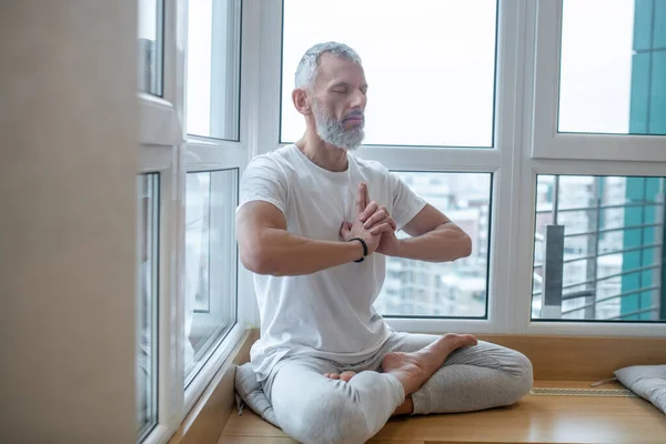 Hombre de pelo gris con una camiseta blanca haciendo yoga y buscando concentrado — Foto de Stock