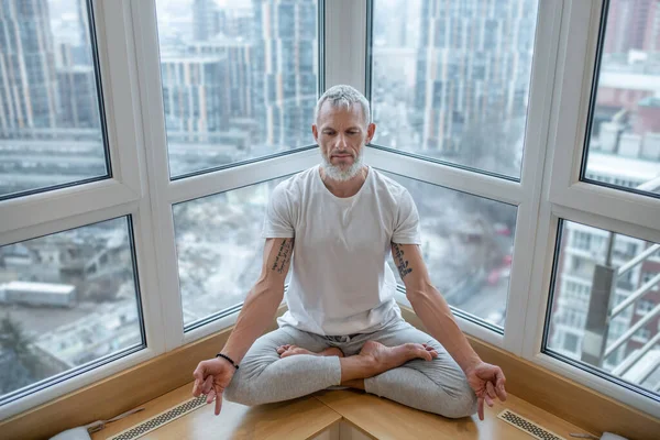 Šedovlasý muž v bílém tričku dělá jógu a vypadá soustředěně — Stock fotografie
