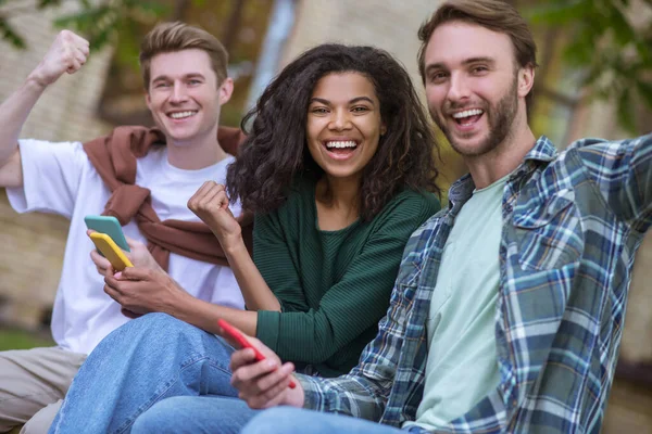 Grupo de jóvenes sentados en el behcn y sosteniendo teléfonos inteligentes en las manos —  Fotos de Stock