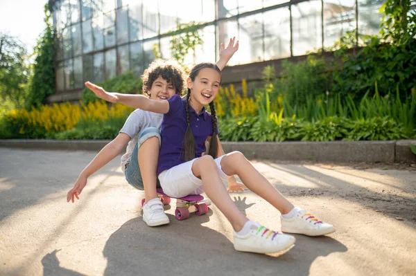 Alegre niño y niña sentado en el monopatín en el parque —  Fotos de Stock