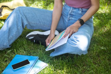 Esmer tenli bir kadın çimenlerde oturup ders çalışıyor.