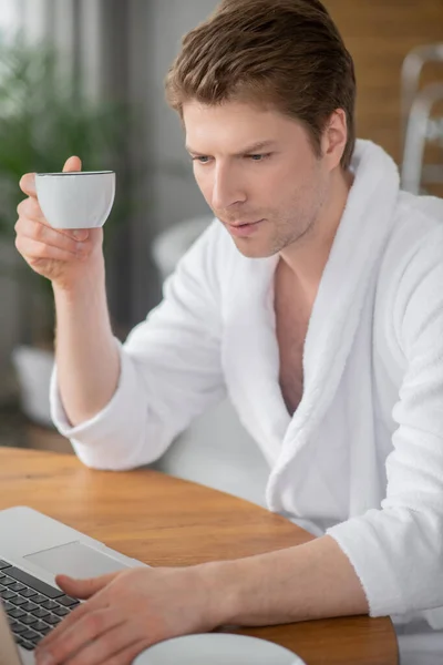 Un hombre guapo en un albornoz blanco sentado en el portátil y pasar tiempo en línea — Foto de Stock