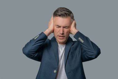 Göz kapakları sarkmış bir adam elleriyle kulaklarını kapatıyor.