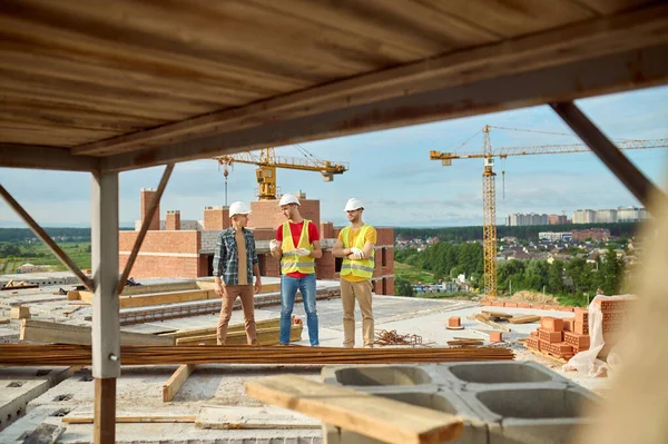 Tři dělníci v těžkých kloboucích vedou rozhovor na staveništi — Stock fotografie