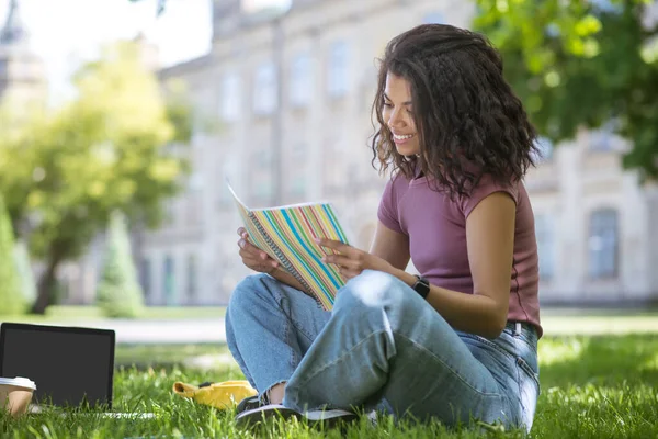 Pembe tişörtlü güzel bir kız parkta çimlerde oturuyor ve ders çalışıyor. — Stok fotoğraf