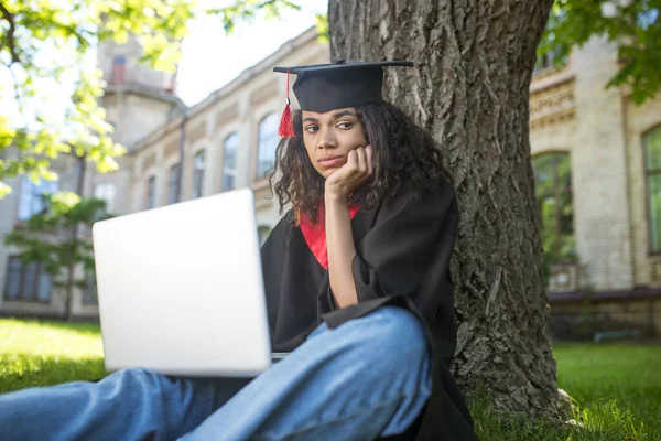 Une fille en robe académique assise sous l'arbre avec un ordinateur portable — Photo