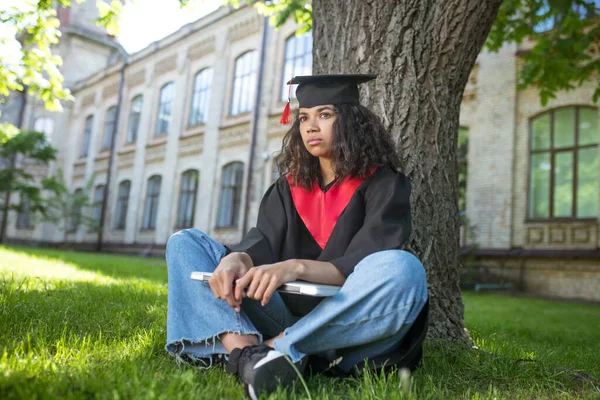 Uma menina de vestido acadêmico sentada debaixo da árvore com um laptop — Fotografia de Stock