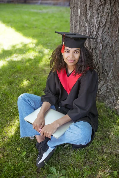 Una chica con bata académica sentada debajo del árbol con un portátil —  Fotos de Stock