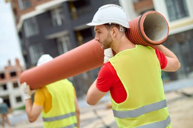 Koruyucu miğferler takmış iki işçi, kıvrımlı bir boru taşıyor.
