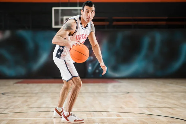 Ung atletisk man i vita sportkläder spelar basket-boll i gymmet — Stockfoto