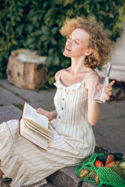 Kızıl kız elinde kitapla evinin yanında oturuyor.