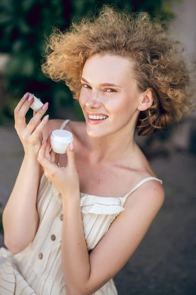 Uma menina de gengibre bonito segurando um frasco de creme e sorrindo — Fotografia de Stock
