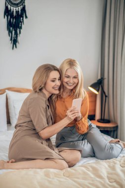 İki gülümseyen sarışın kadın yatakta oturuyor ve akıllı telefonda selfie çekiyorlar.
