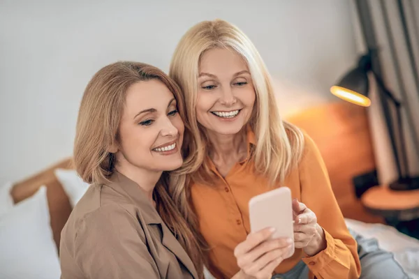 Dos mujeres rubias sonrientes sentadas en la cama y haciendo selfie en un smartphone — Foto de Stock