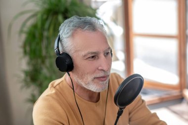 Siyah kulaklıklı, gri saçlı bir adam mikrofonla konuşuyor ve meşgul görünüyor.