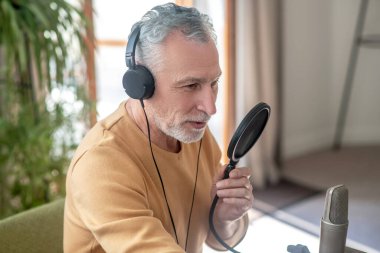 Siyah kulaklıklı, gri saçlı bir adam mikrofonla konuşuyor ve meşgul görünüyor.